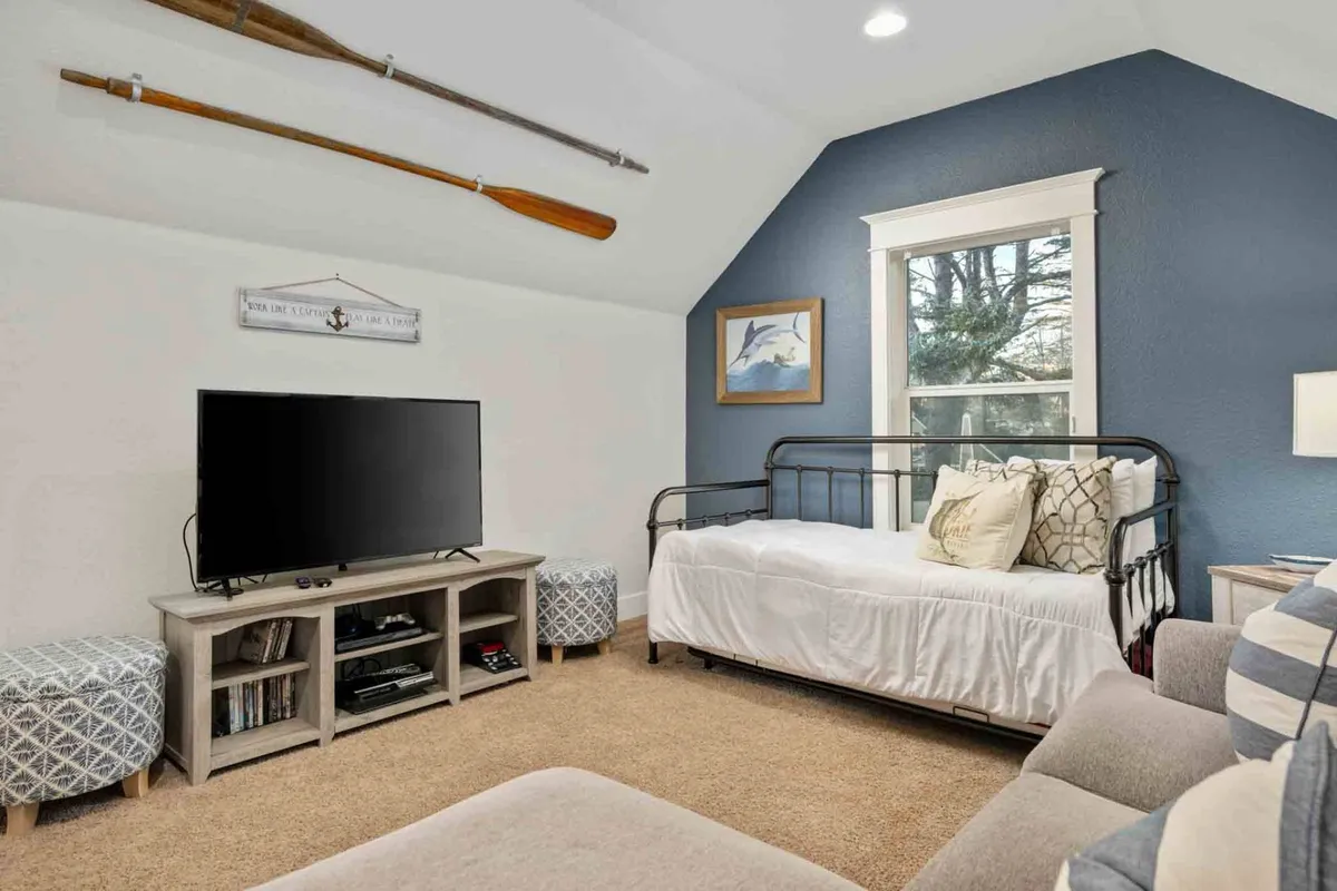 This image showcases a cozy, playful loft area featuring a daybed with decorative pillows, a television on a media console, framed artwork, two decorative ottomans, and wooden rowing oars on the wall. The room is bathed in natural light and designed for relaxation, socialization, and fun.