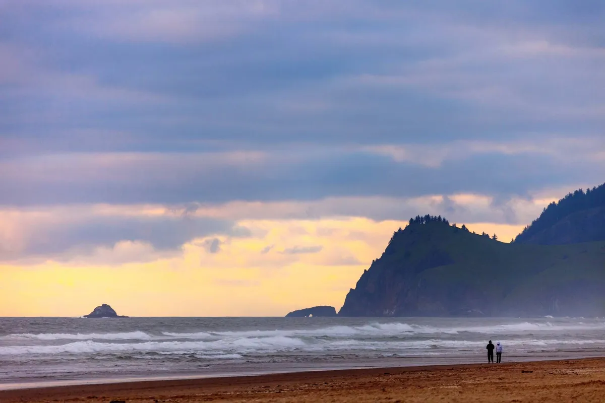 Scenic ocean waves roll gently toward the sandy beach, with distant cliffs in the background. A couple strolls along the shoreline, highlighting the spaciousness and natural beauty of the coast.