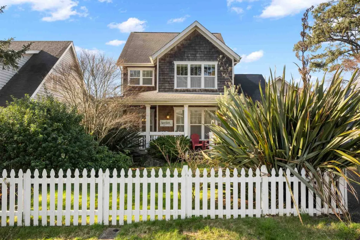 The exterior of the Storybook Beach Cottage is depicted, with a charming white picket fence, a front porch featuring red chairs, and lush greenery surrounding the property. The scene is cozy and welcoming, suggesting a safe and enjoyable space for guests and their pets to relax outdoors.
