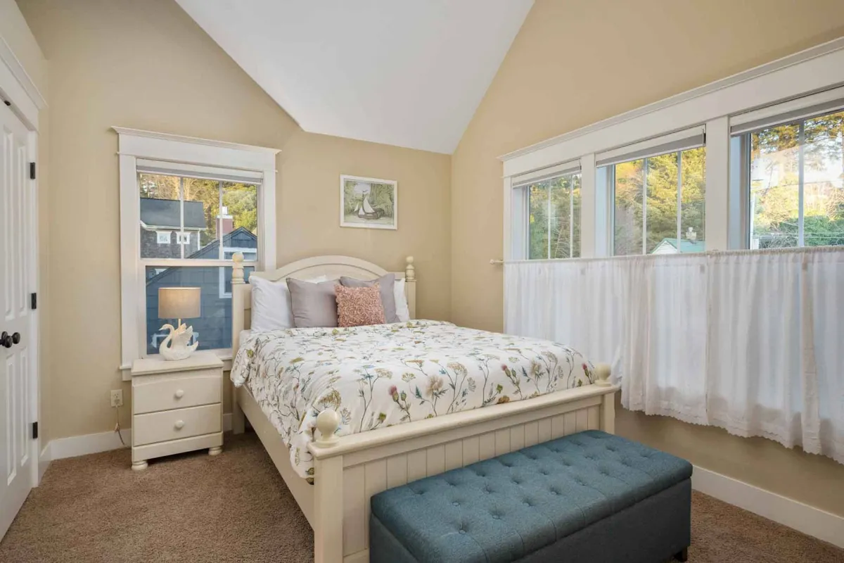 A serene bedroom featuring a bed with floral bedding, large windows letting in natural light, decorative images on the walls, a storage nightstand, and a blue ottoman. The room feels tranquil, ideal for restful mornings and quiet reading.