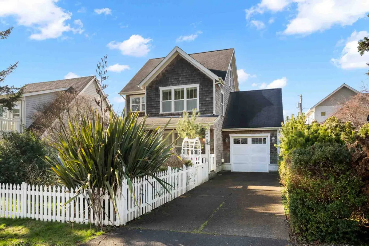 A cheerful view of the house with a charming gabled roof and a classic white picket fence, surrounded by vibrant greenery in the front yard. The porch and garden evoke a sense of leisurely outdoor living.