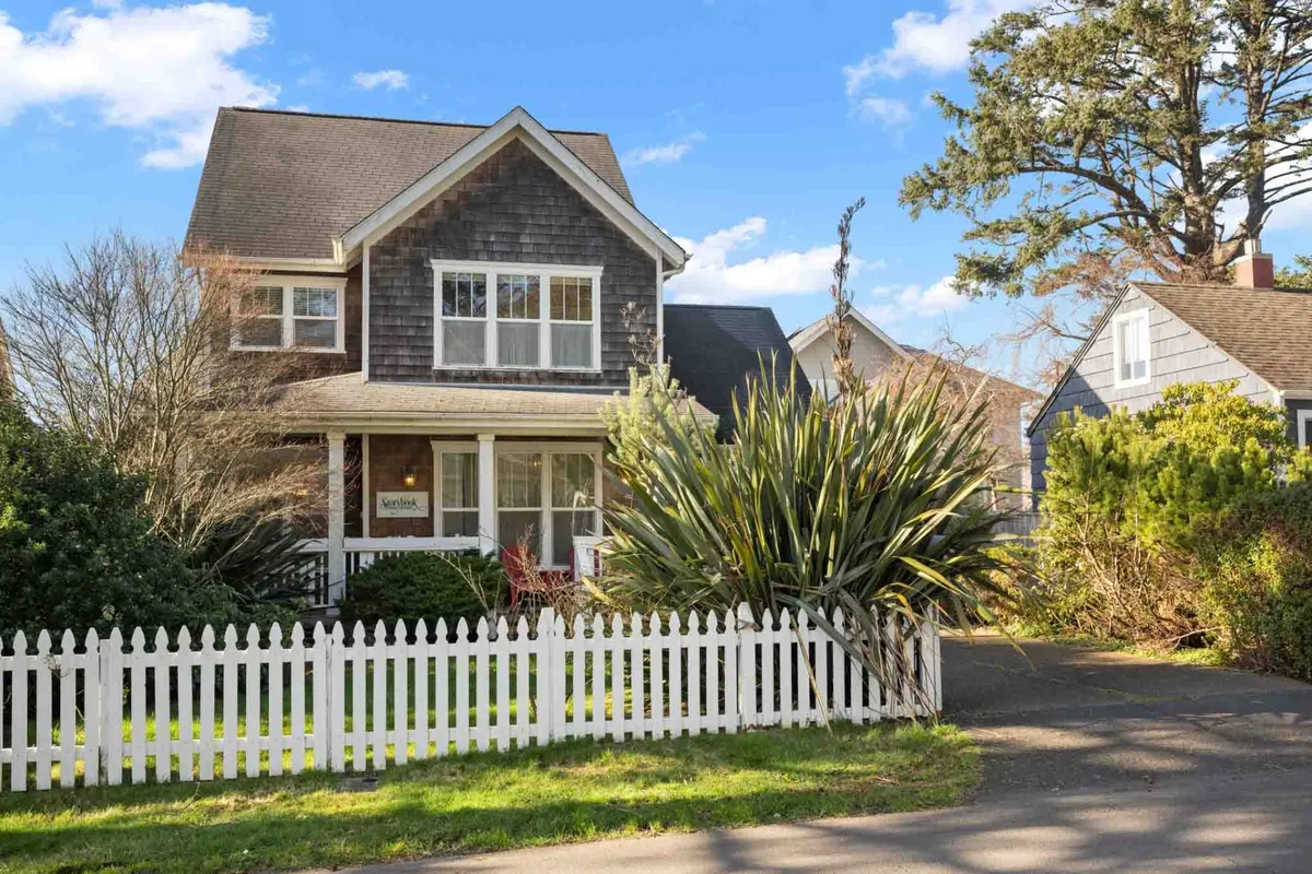 A two-story house with a welcoming front porch sits behind a classic white picket fence, surrounded by lush greenery. The porch features seating, inviting relaxation or group gatherings. The overall setting suggests a family-friendly environment ideal for both privacy and connection.