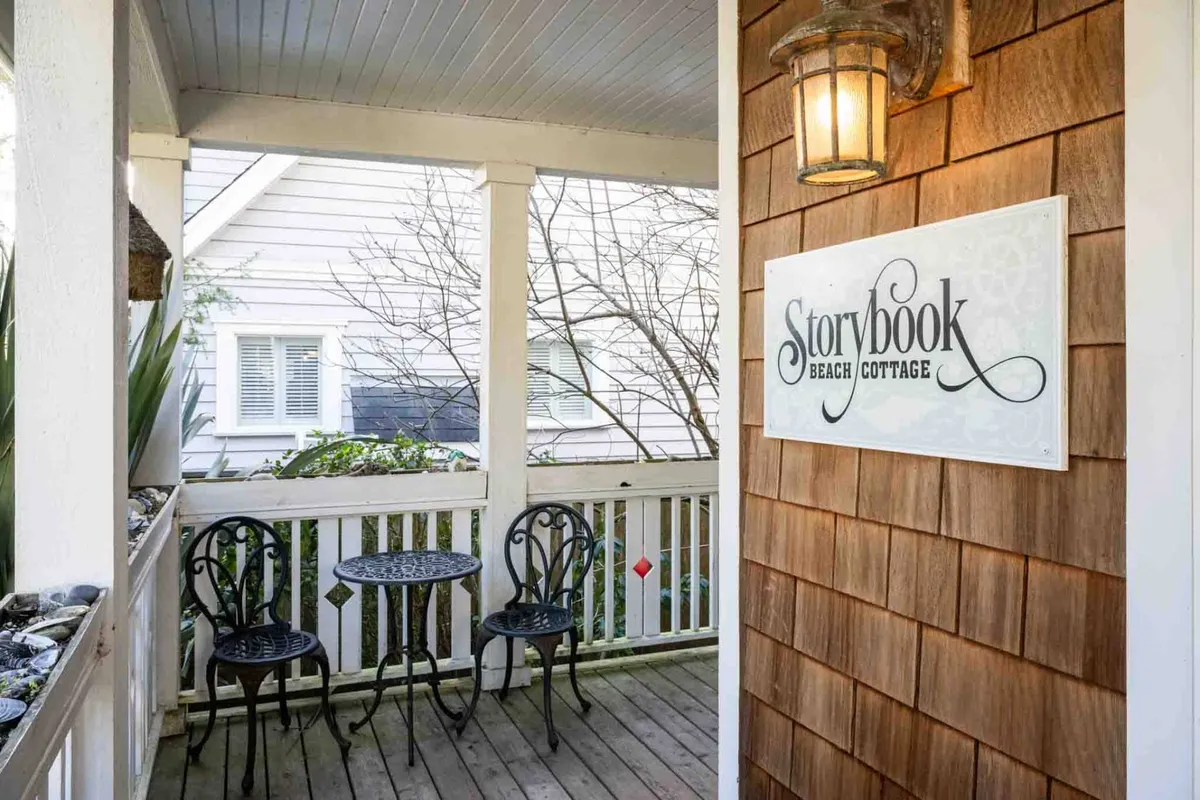 The charming exterior of Storybook Beach Cottage, with a visible sign, metal table and chairs, and classic wooden siding. The cozy porch scene is ideal for relaxing after a day of seaside fun.