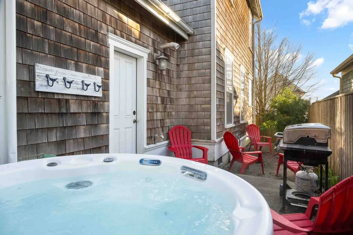 This image showcases a cozy backyard setup featuring a hot tub, bright red chairs, a grill, and a convenient door leading back inside. The space is clearly designed for both relaxation and social gatherings, with ample room for pets and children to play.