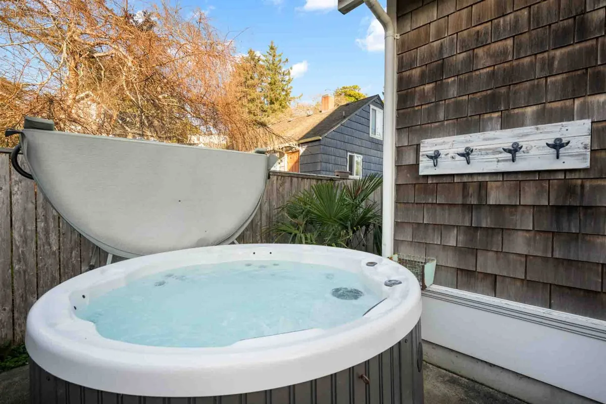 A bubbling hot tub sits nestled amid lush greenery and wooden house siding, with coat hooks and outdoor space for entertaining or relaxing. The setting is perfect for evening soaks or social time with guests.